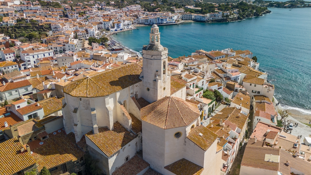 Vista aérea del casco antiguo de Cadaqués, un pintoresco pueblo de la Costa Brava, con tejados rojizos, la iglesia de Santa María en primer plano y el mar Mediterráneo al fondo.