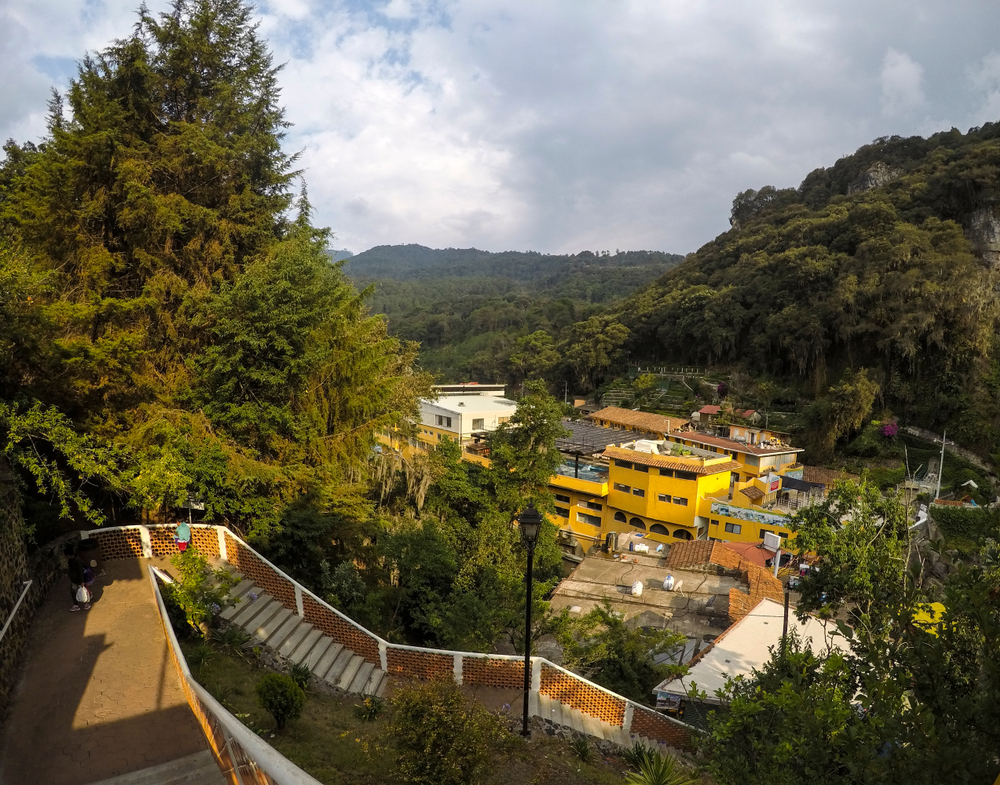 Vista panorámica de Chignahuapan, Puebla, con casas de colores brillantes, entre ellas una edificación amarilla destacada, rodeadas de frondosos árboles de pino y montañas boscosas. En primer plano se observa una escalinata con barandales de ladrillo y una persona descendiendo, mientras el cielo parcialmente nublado resalta la atmósfera fresca y natural del pueblo.