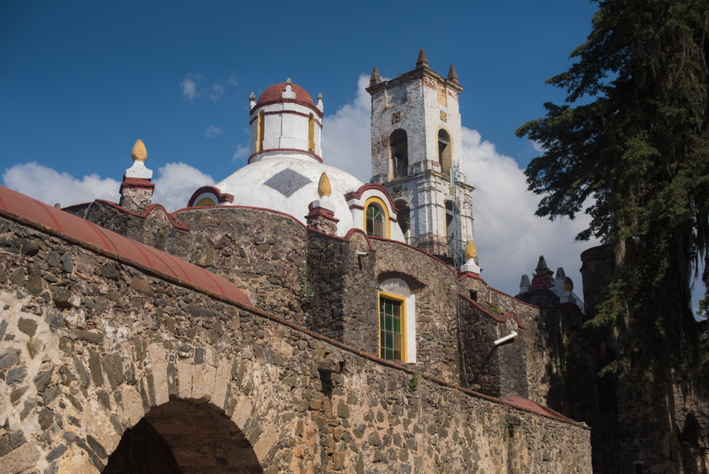 Fotografía del Ex Convento de San Juan Bautista en Huasca de Ocampo, Hidalgo, México. Se aprecia su arquitectura colonial de piedra volcánica, con cúpula blanca decorada en rojo y una torre campanario desgastada por el tiempo. El cielo azul y la vegetación que rodea el conjunto resaltan el ambiente histórico y pintoresco del primer Pueblo Mágico de México.
