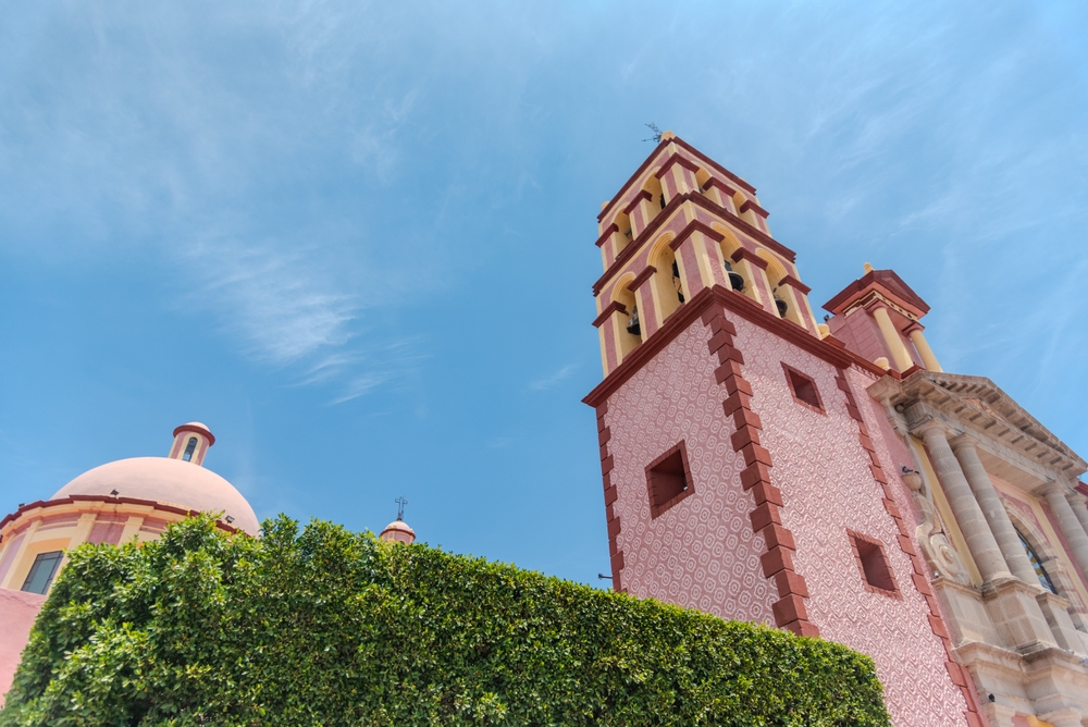 Fotografía de la Parroquia de Santa María de la Asunción en Tequisquiapan, Querétaro. Se observa su torre principal de color rosa con detalles geométricos, cúpula redonda y fachada barroca bajo un cielo azul claro. Al frente, un seto verde enmarca el templo, símbolo arquitectónico del centro histórico del Pueblo Mágico.