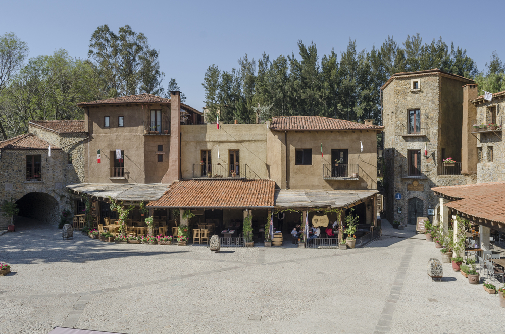 Plaza principal de Val’Quirico, un desarrollo turístico en Tlaxcala, México, con arquitectura inspirada en pueblos medievales europeos. Se observan edificaciones de piedra y adobe con techos de teja, balcones decorados con banderas mexicanas, y terrazas con sillas de madera rodeadas de macetas florales. Al fondo, árboles altos completan el entorno pintoresco y tranquilo del lugar.