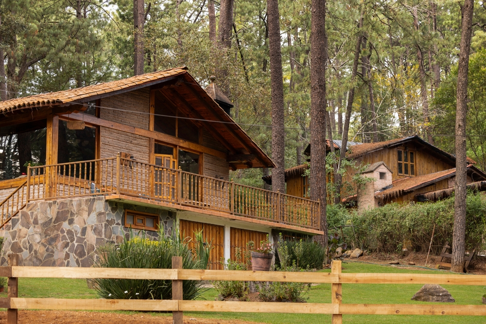 Cabañas rústicas construidas con piedra y madera entre un bosque de pinos, rodeadas de naturaleza en una zona montañosa cercana a Ciudad de México. La escena muestra una cabaña principal con terraza y barandales de madera, césped bien cuidado y una cerca de madera en primer plano, ideal para escapadas tranquilas en contacto con la naturaleza.