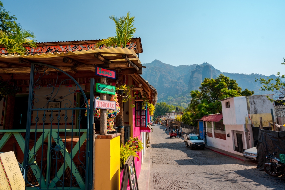 Calle empedrada en Tepoztlán, México, con fachadas coloridas, vegetación tropical y un restaurante decorado con carteles artesanales que anuncian comida típica como sopes y guacamole. Al fondo, se alzan las montañas del Tepozteco bajo un cielo despejado. El entorno refleja la mezcla de cultura tradicional y naturaleza de este pueblo mágico.