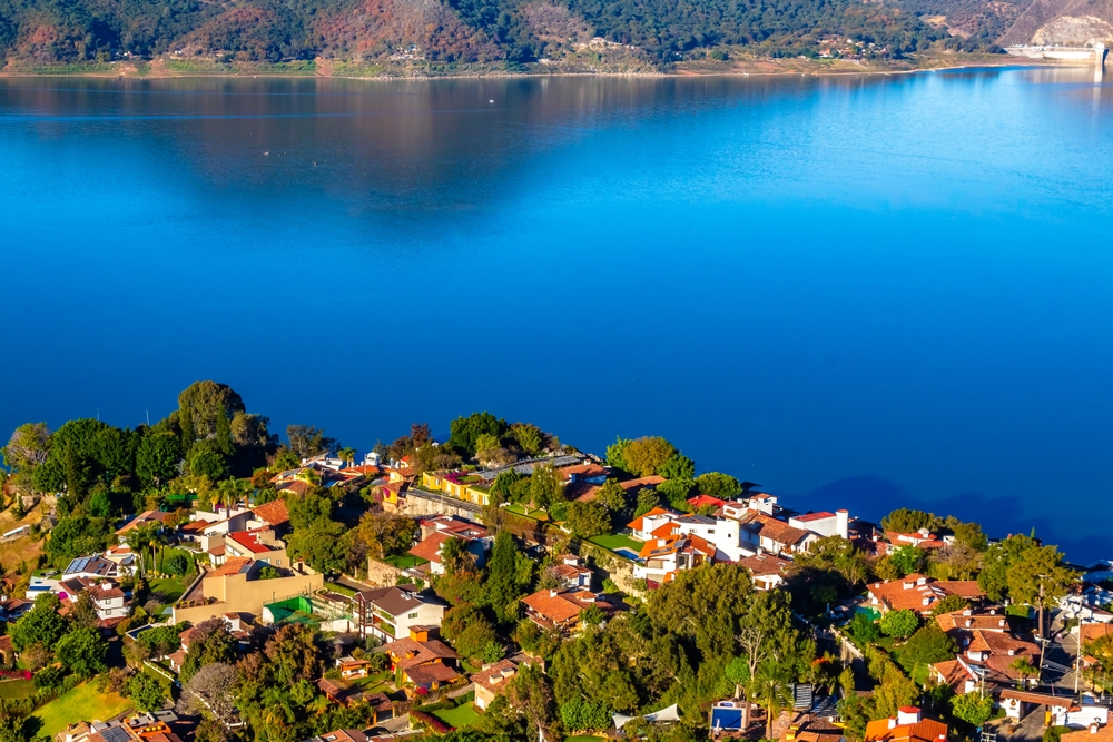 Fotografía panorámica de Valle de Bravo, México, con casas coloridas entre vegetación frondosa en primer plano, junto al lago con aguas azules intensas que reflejan el cielo. Al fondo, se observan montañas arboladas y la serenidad del paisaje natural. Es un destino popular para escapadas de naturaleza, deportes acuáticos y turismo rural.