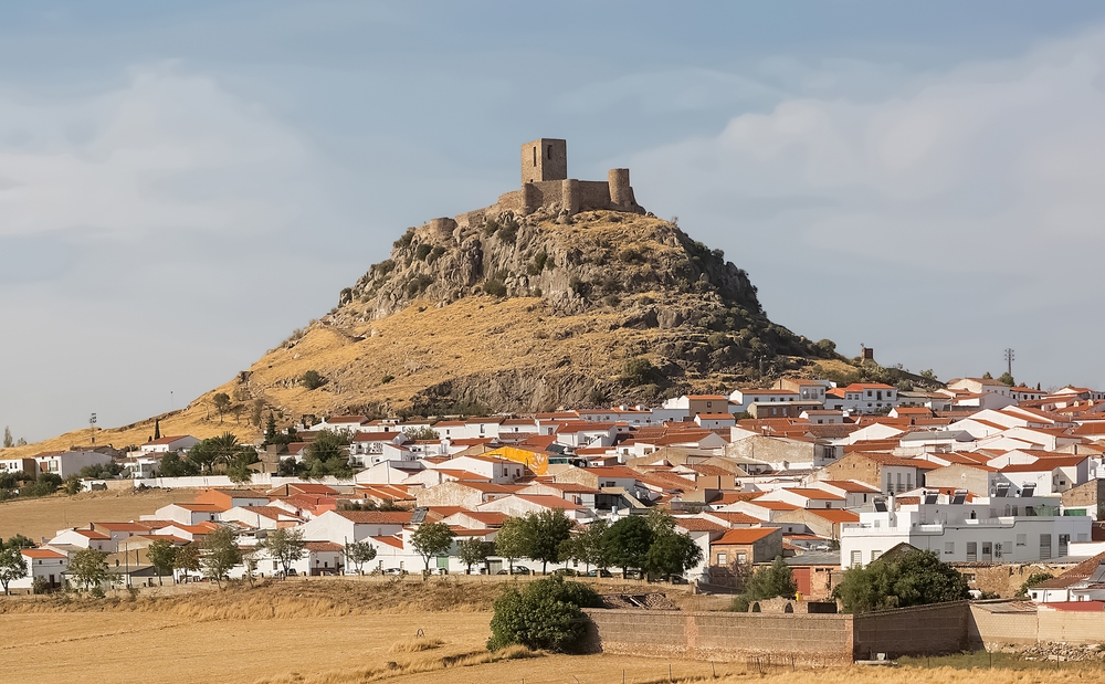 Vista panorámica del pueblo de Bélmez de la Moraleda, en Jaén, España, con casas de tejados rojizos y fachadas blancas. Al fondo se eleva un cerro rocoso coronado por el Castillo de Bélmez, una fortificación medieval en ruinas que domina el paisaje. El entorno árido y dorado refleja el clima seco de la región. Este pueblo es conocido por las supuestas apariciones paranormales en una vivienda local, conocidas como las "Caras de Bélmez".