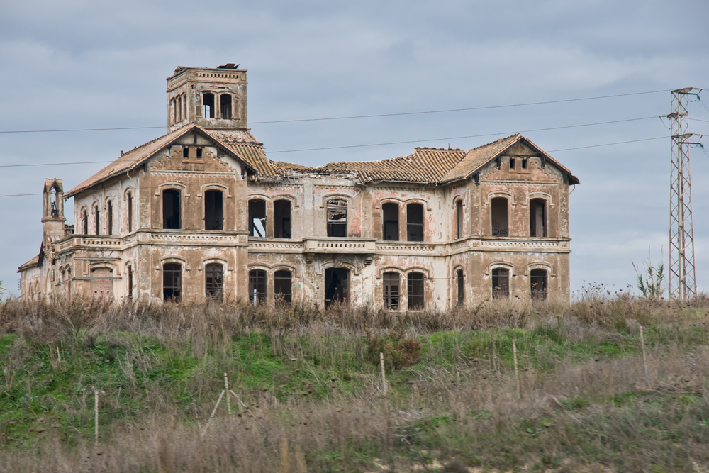 Fotografía del Cortijo Jurado, una mansión abandonada y de aspecto tenebroso ubicada en Málaga, España. El edificio de estilo neoclásico muestra su fachada deteriorada, con paredes desgastadas, ventanas rotas y tejado dañado. Está rodeado de vegetación silvestre y hierba alta, bajo un cielo nublado que refuerza su atmósfera misteriosa.