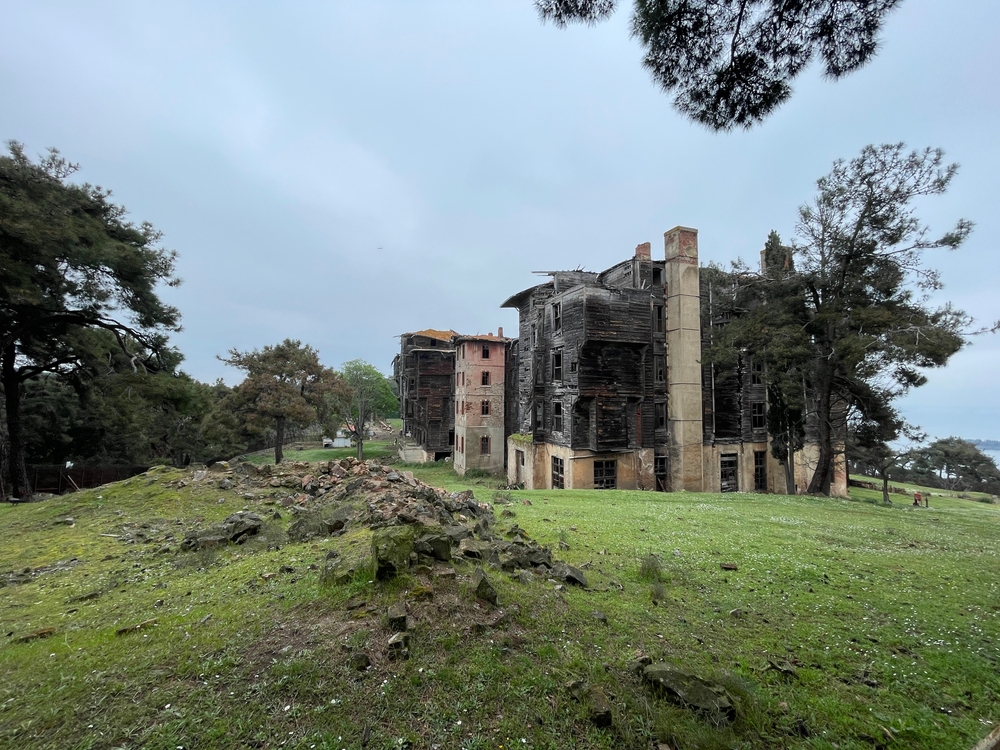 Fotografía del antiguo Sanatorio de Cesuras, ubicado en un claro rodeado de árboles en Galicia, España. El edificio principal, de varios pisos y visiblemente deteriorado, combina secciones de ladrillo con estructuras de madera quemada y desprendida. La construcción está parcialmente en ruinas, rodeada de césped verde y restos de piedra, bajo un cielo nublado que acentúa su atmósfera melancólica y abandonada.