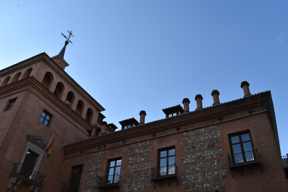 Fotografía de la Casa de las Siete Chimeneas en Madrid, España, tomada desde un ángulo bajo. El edificio, de ladrillo rojo con detalles en piedra y balcones de hierro forjado, destaca por sus siete chimeneas visibles en el tejado. A la izquierda, se aprecia una torre con un veleta y una bandera de España ondeando sobre un balcón. El cielo despejado y azul contrasta con la arquitectura histórica del inmueble.