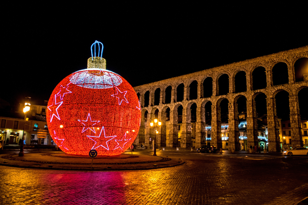Vista nocturna del Acueducto de Segovia iluminado, con una gran esfera navideña roja decorada con estrellas frente a él. La esfera está iluminada con luces LED rojas, blancas y azules, simulando un adorno navideño gigante. Las calles adoquinadas reflejan las luces, mientras que edificios y farolas completan el ambiente festivo