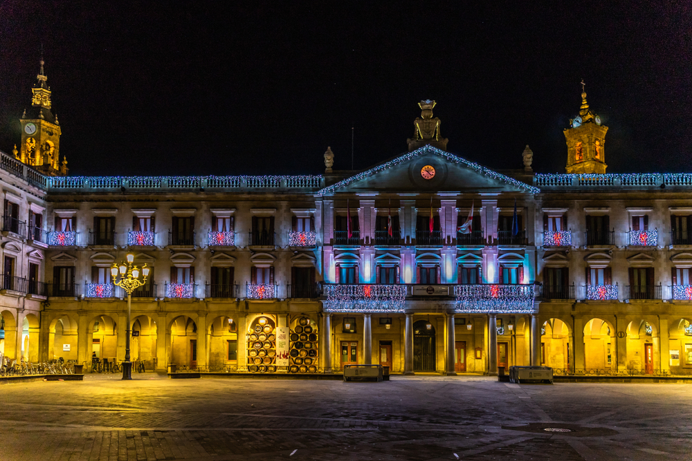 Plaza iluminada en Vitoria-Gasteiz durante la Navidad. Vista nocturna de la fachada del Ayuntamiento adornada con luces navideñas en tonos azules, blancos y rojos. El edificio histórico de tres pisos destaca con balcones decorados y una gran figura en la azotea. A ambos lados se elevan torres con relojes y cúpulas iluminadas. La plaza está vacía, con un suelo adoquinado y una farola antigua al frente. Ambiente festivo y elegante.