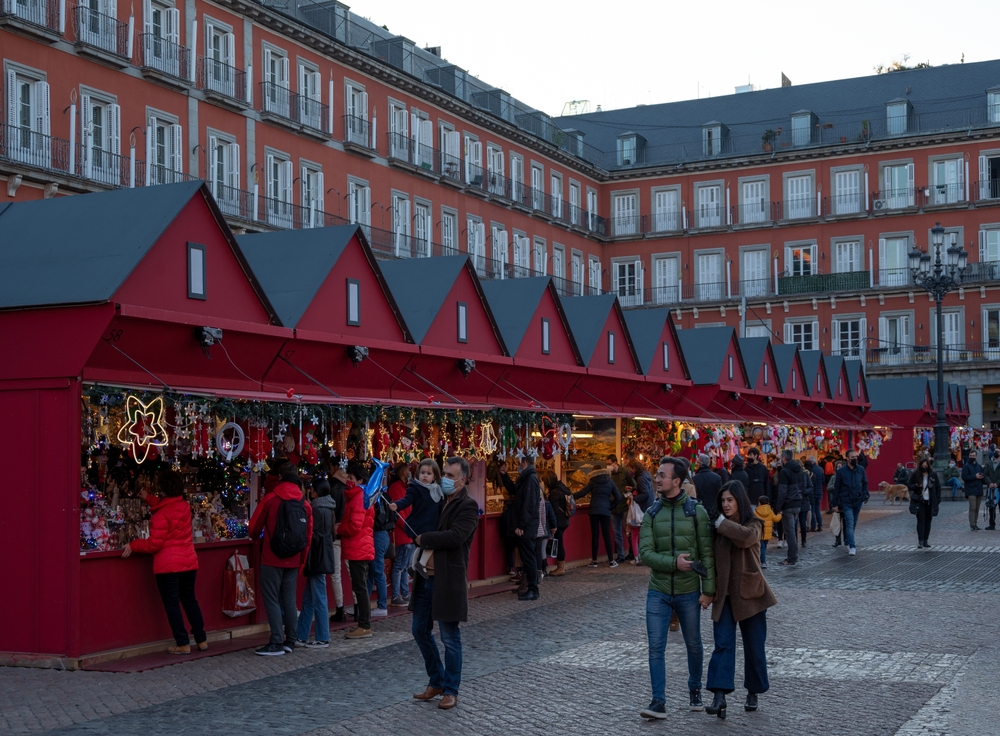 Mercado navideño en la Plaza Mayor de Madrid, con casetas rojas alineadas y decoradas con luces y adornos. Varias personas pasean, compran y observan los puestos en un ambiente festivo. Al fondo, los edificios históricos de la plaza completan la escena invernal.
